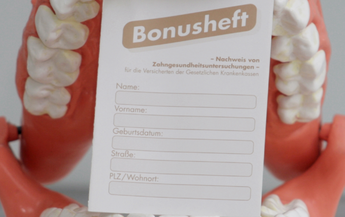 Ein Plastikgebiss hält ein Bonusheft zwischen den Zähnen.